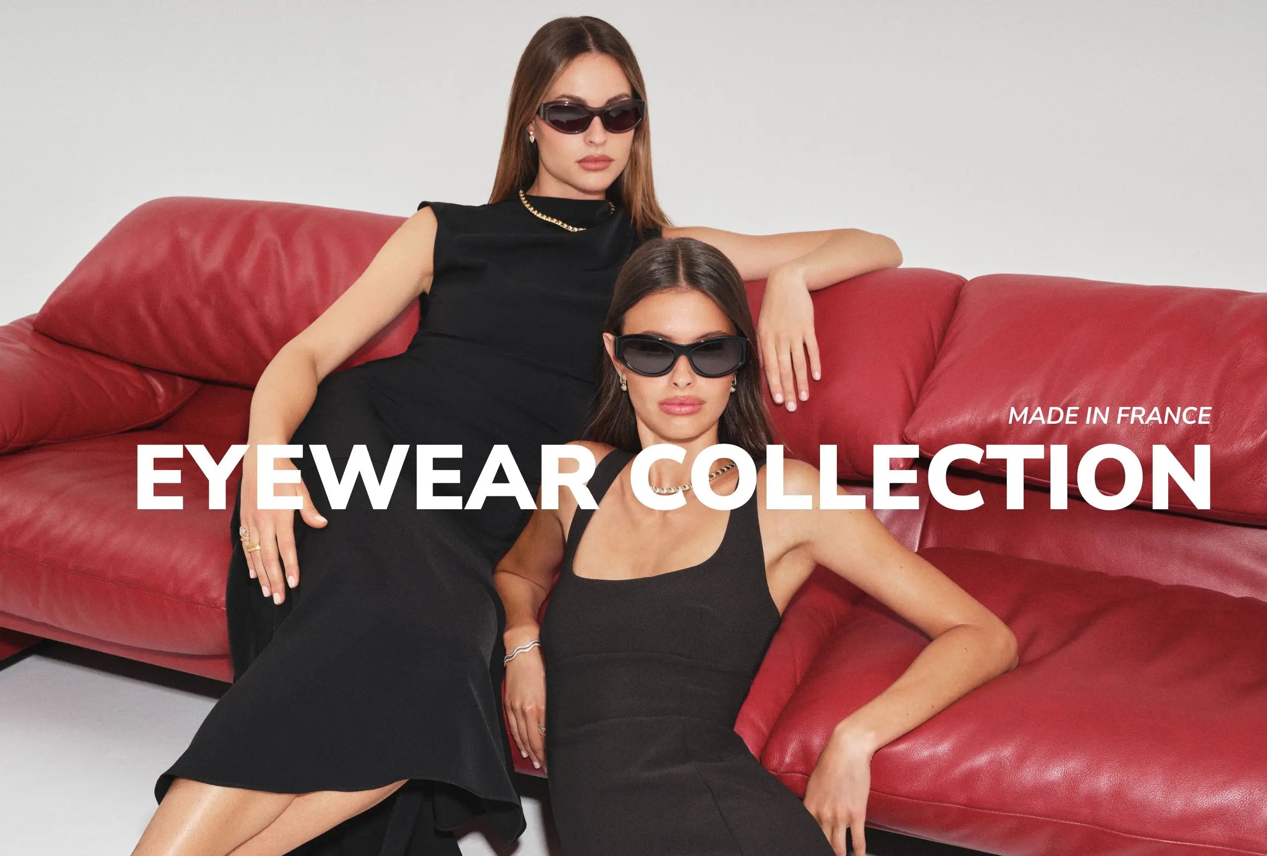 Two women sitting on a red couch wearing black outfits and sunglasses.